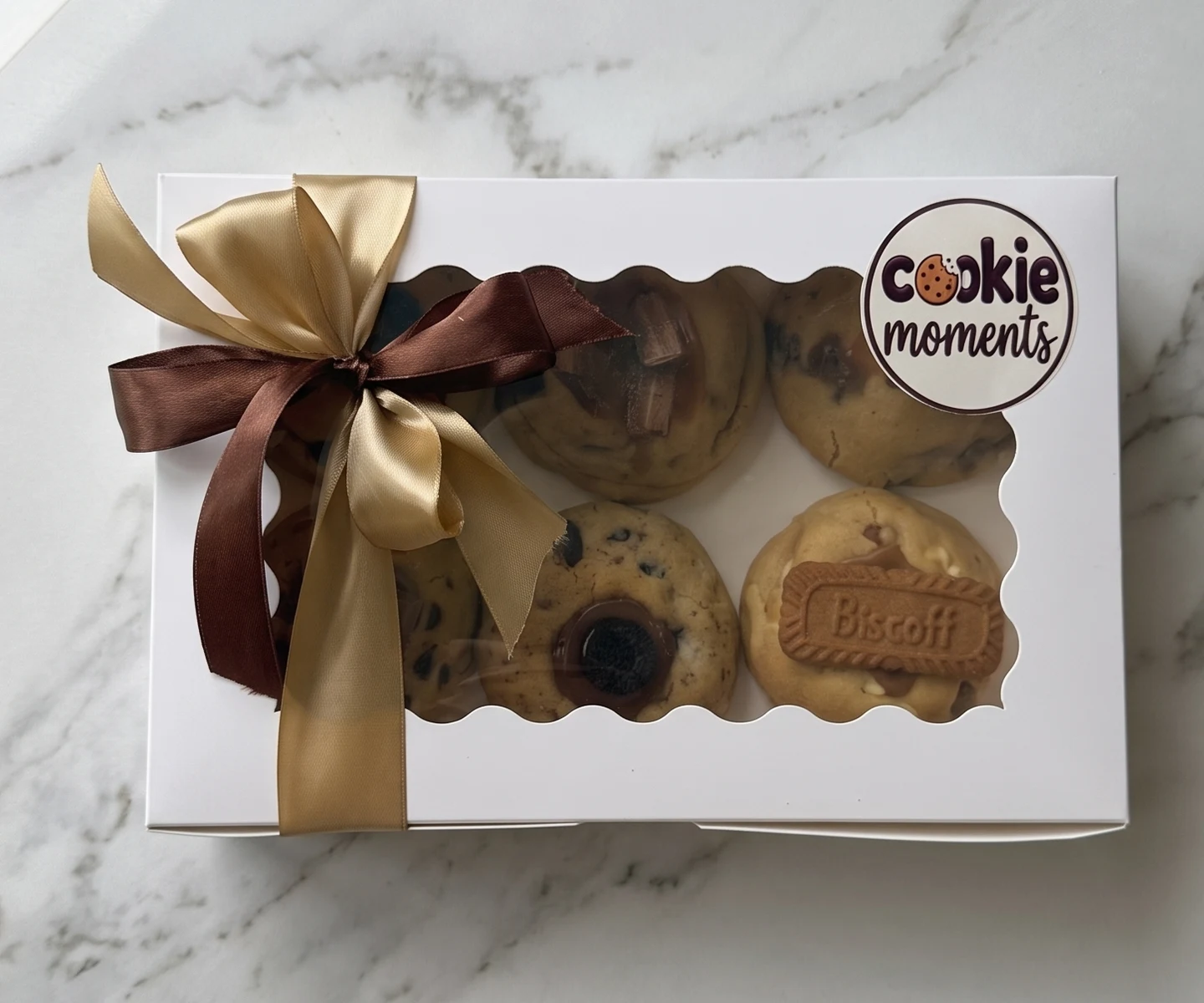 A Cookie Moments box of six on a white marble bench, tied with brown and gold ribbon, Biscoff, Oreo and caramel cookies visible through the scalloped window